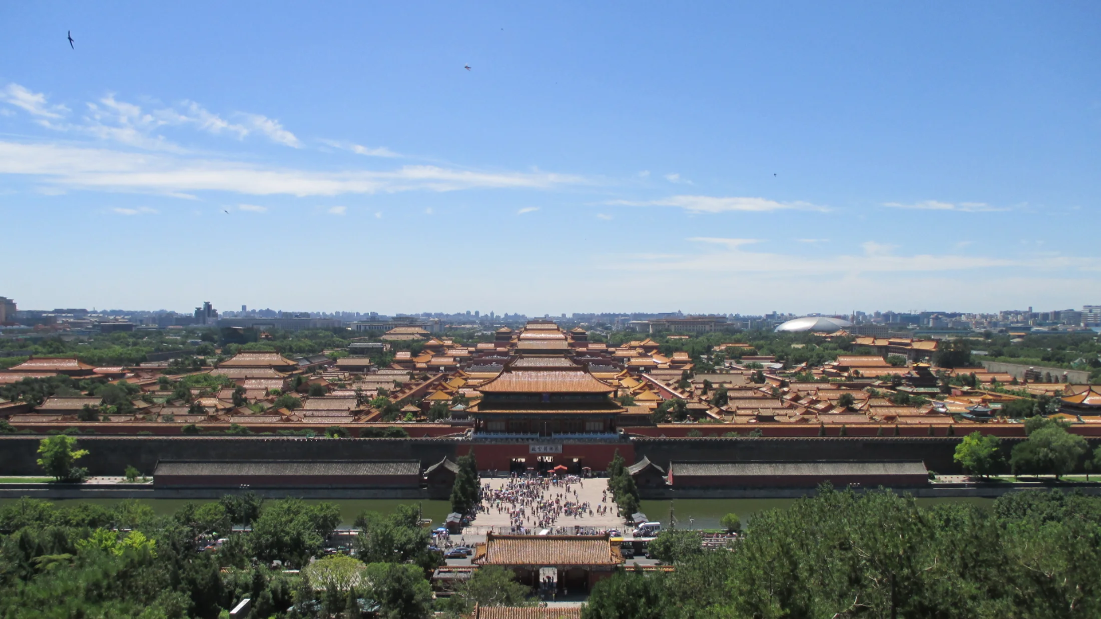 The Forbidden City's long sequence of red walls and golden roofs extends south from Jingshan under a clear blue sky, with contemporary Beijing visible beyond.