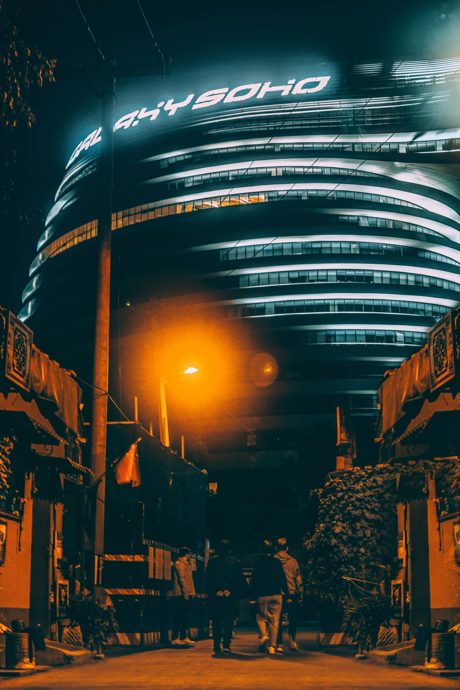 People walk through a low-rise Beijing lane at night beneath the illuminated curved facade of Galaxy SOHO.