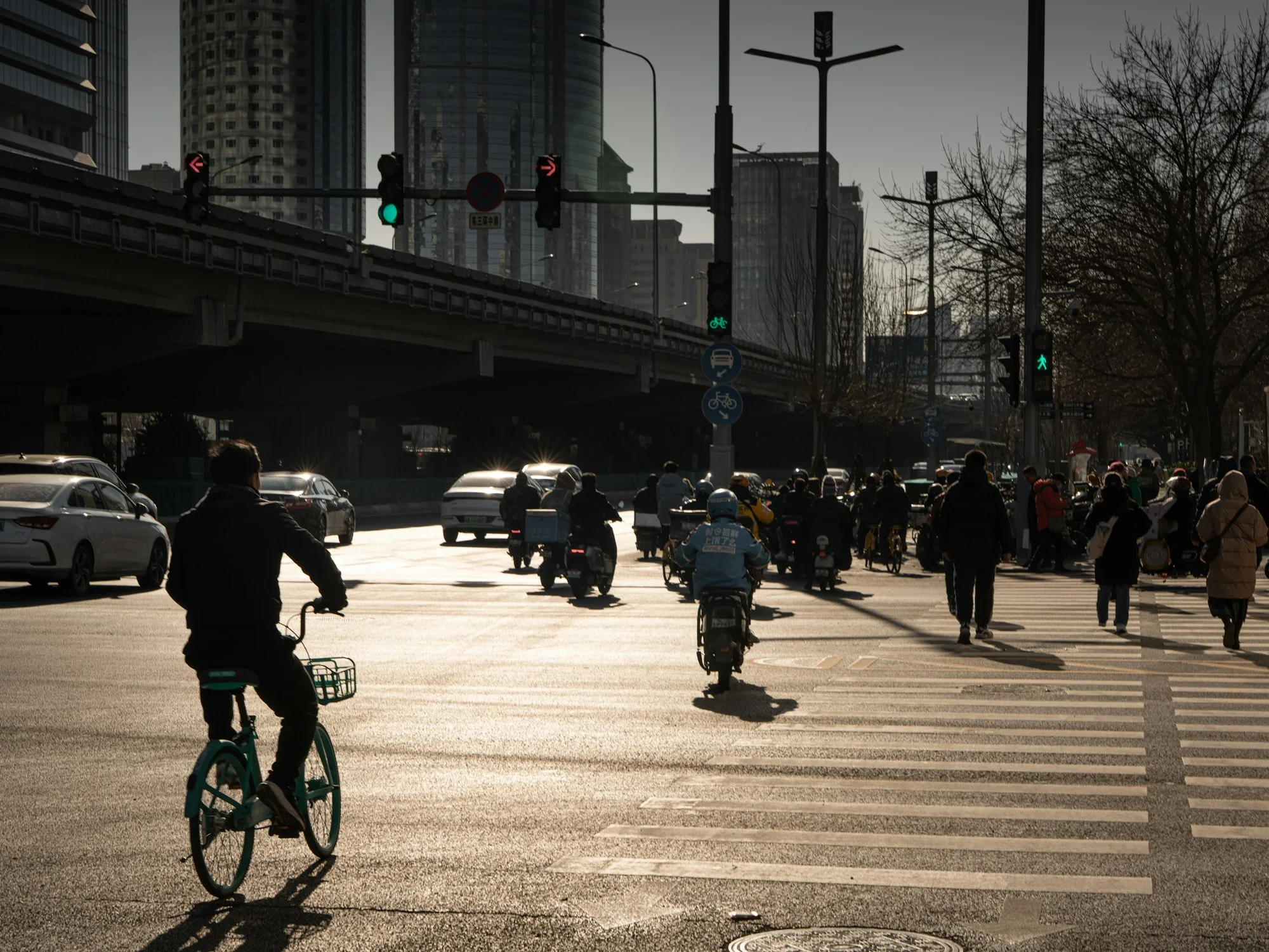 Cyclists, delivery riders, pedestrians and cars cross a broad intersection beneath office towers and elevated roads in Beijing's Guomao district.