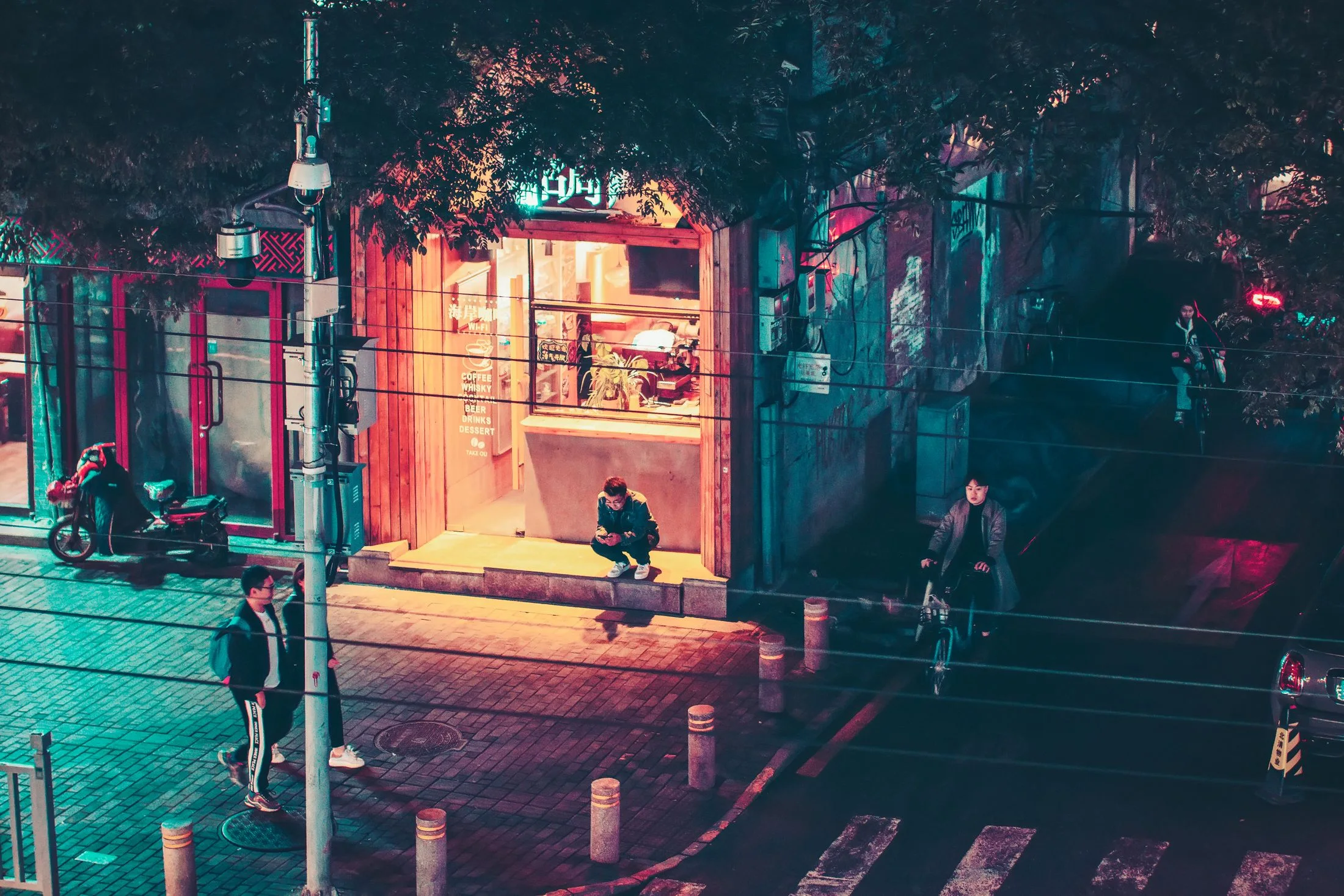 People, a bicycle and a scooter occupy a Beijing neighborhood street beside a small café glowing at night.