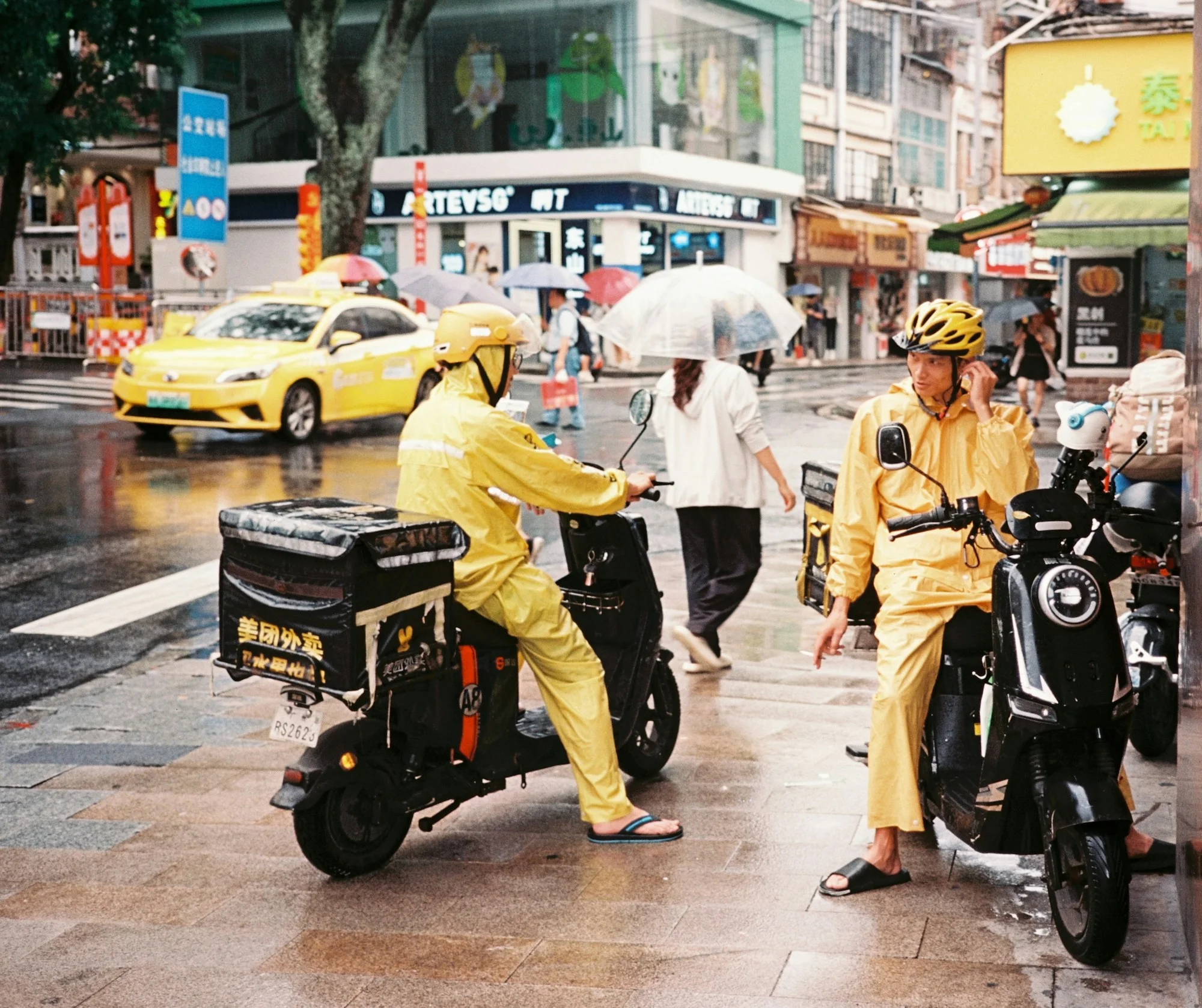 Two food-delivery riders in yellow rainwear wait on a wet Guangzhou street beside a taxi and passing pedestrians.