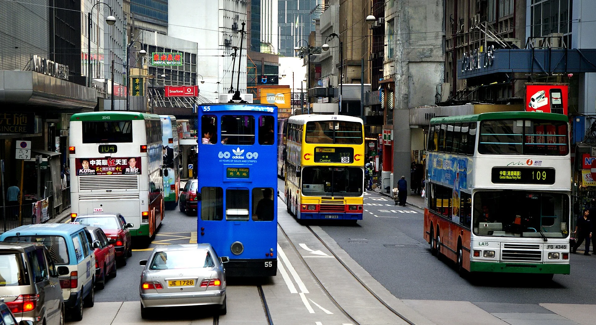 A Hong Kong street layers a double-decker tram, bus, minibus, taxis and dense shop signs.