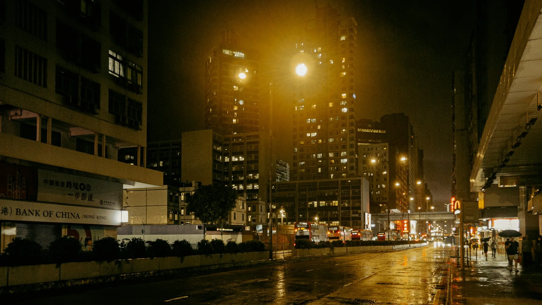 Buses and pedestrians move through a rain-wet Hong Kong street beneath dense residential towers and warm streetlights.