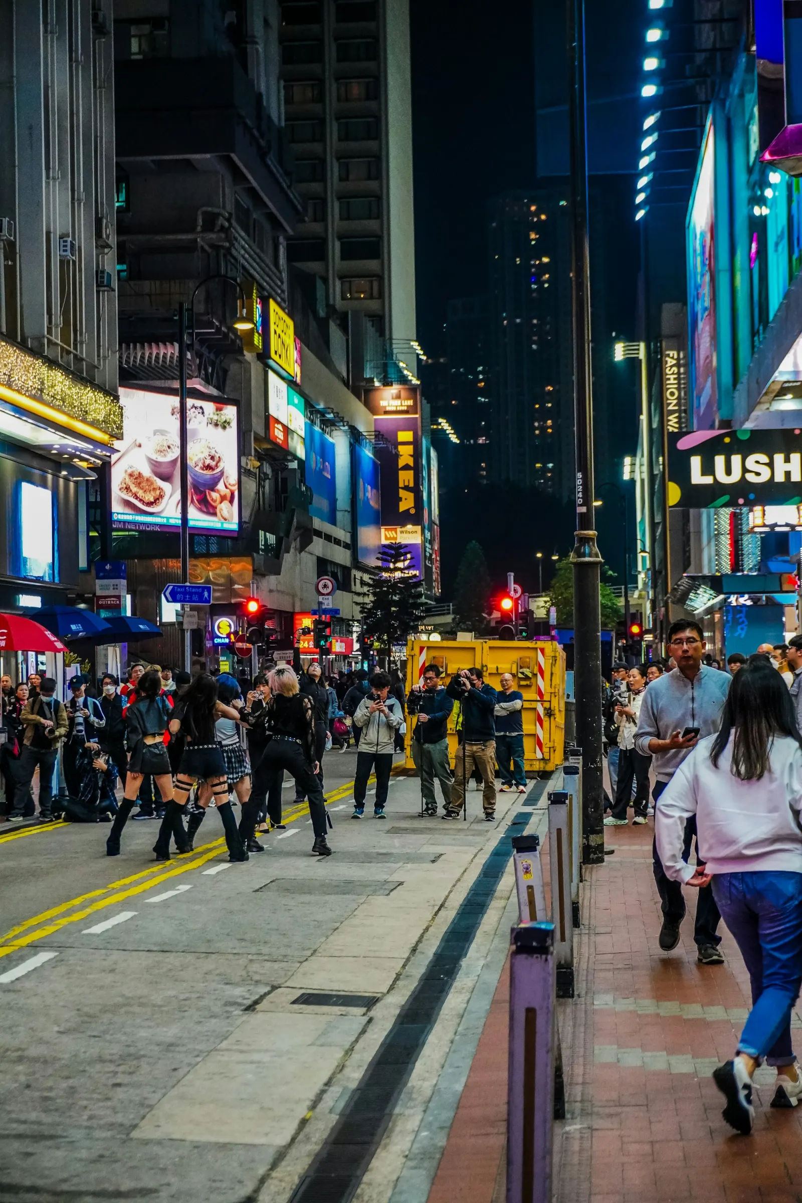 A young crowd watches a live dance performance on a neon-lit Wan Chai street in Hong Kong at night.