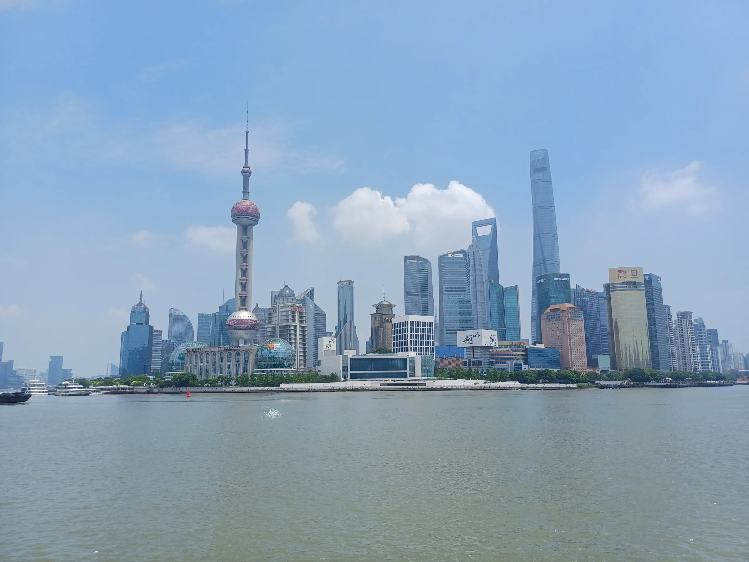 Lujiazui's financial-district skyline rises across the Huangpu River from the Bund under a bright blue sky with moving boats in the foreground.