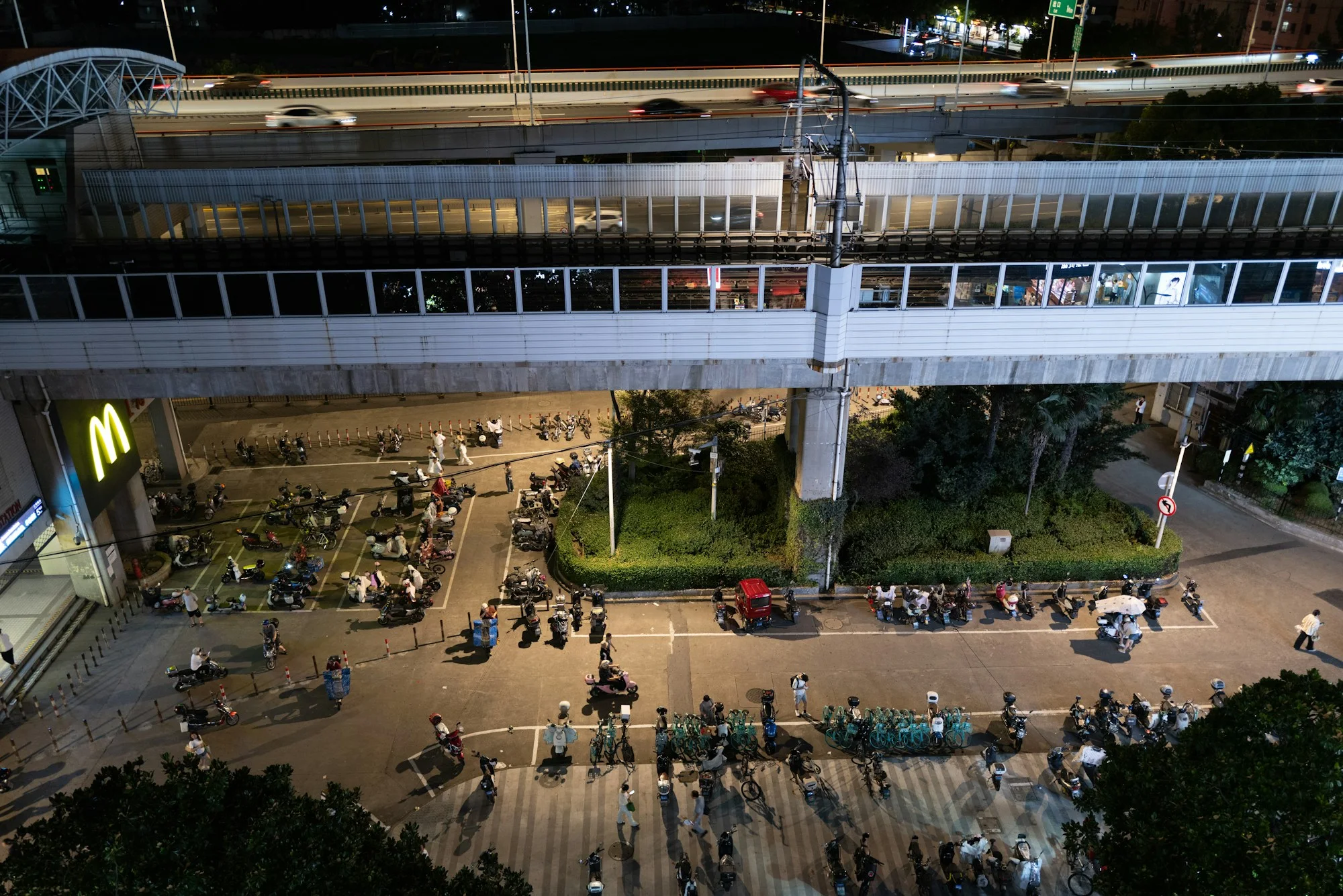 At night in Shanghai, cars move on an elevated road above a train while cyclists, scooters and pedestrians gather at street level.