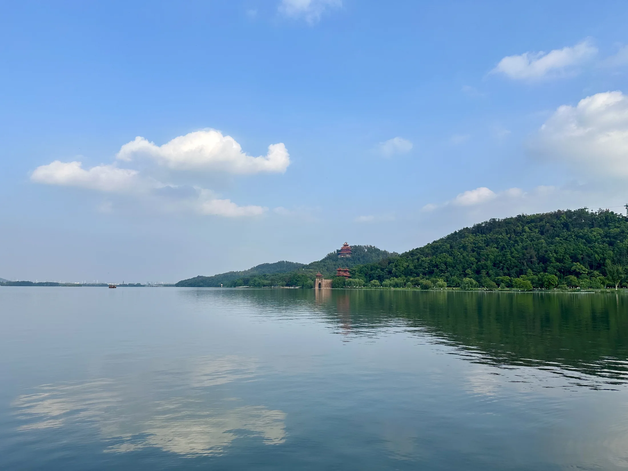 East Lake stretches toward the wooded slopes of Mount Mo under blue sky and white clouds, with a small lakeside structure reflected in calm water.