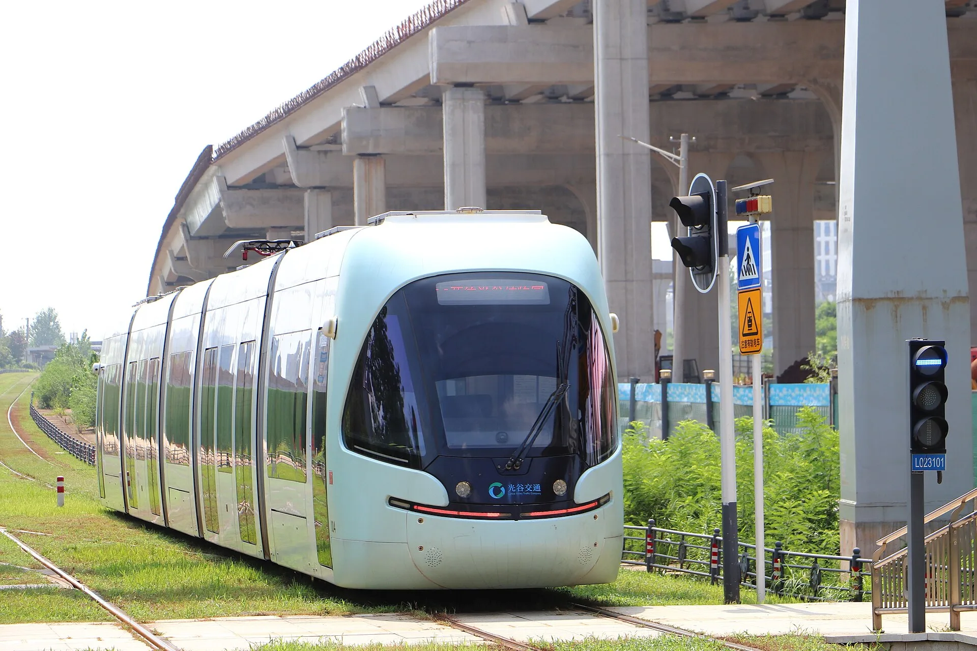 A pale green Optics Valley tram turns through a sunlit Wuhan junction beneath elevated infrastructure.