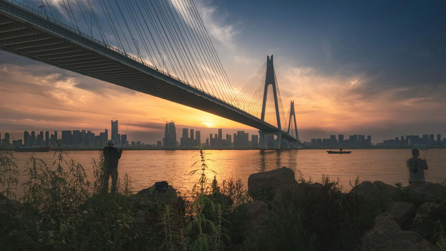 A cable-stayed bridge crosses the Yangtze at sunset in Wuhan while two people fish from the riverbank and the skyline stretches beyond.