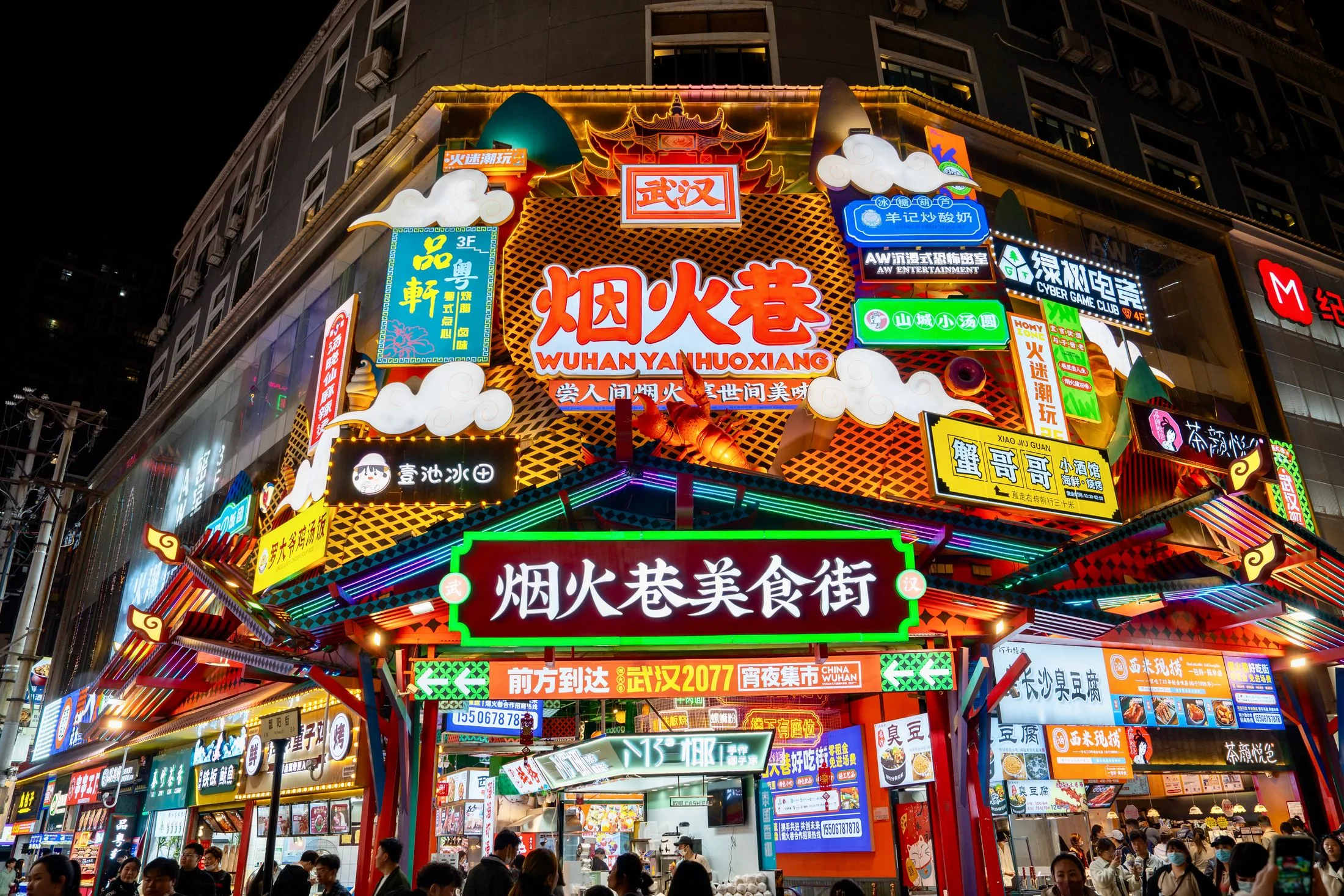 A crowd gathers below dense illuminated food signs at Wuhan's Yanhuoxiang night market.