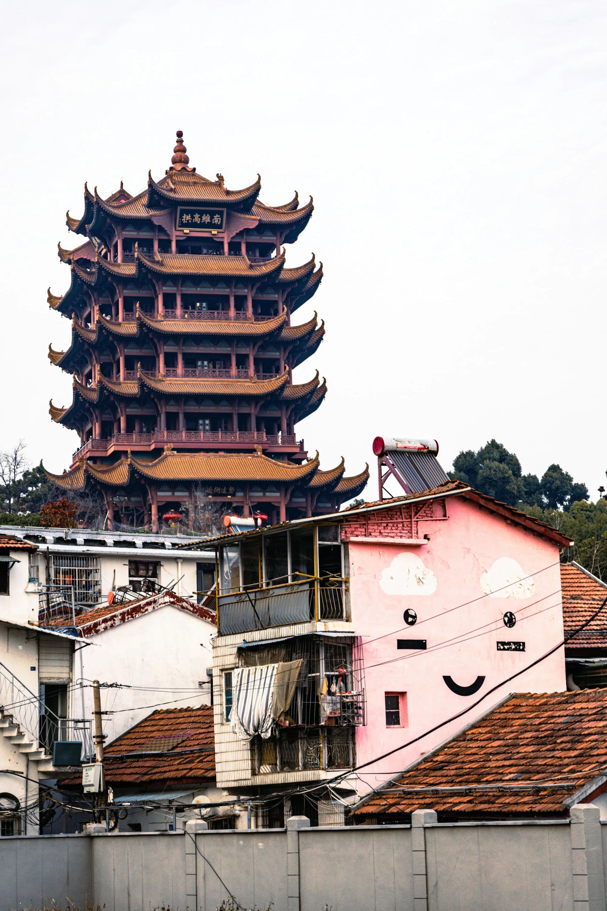 Yellow Crane Tower rises behind closely packed Wuhan homes, tiled roofs, utility wires and a pink wall painted with a smiling face.