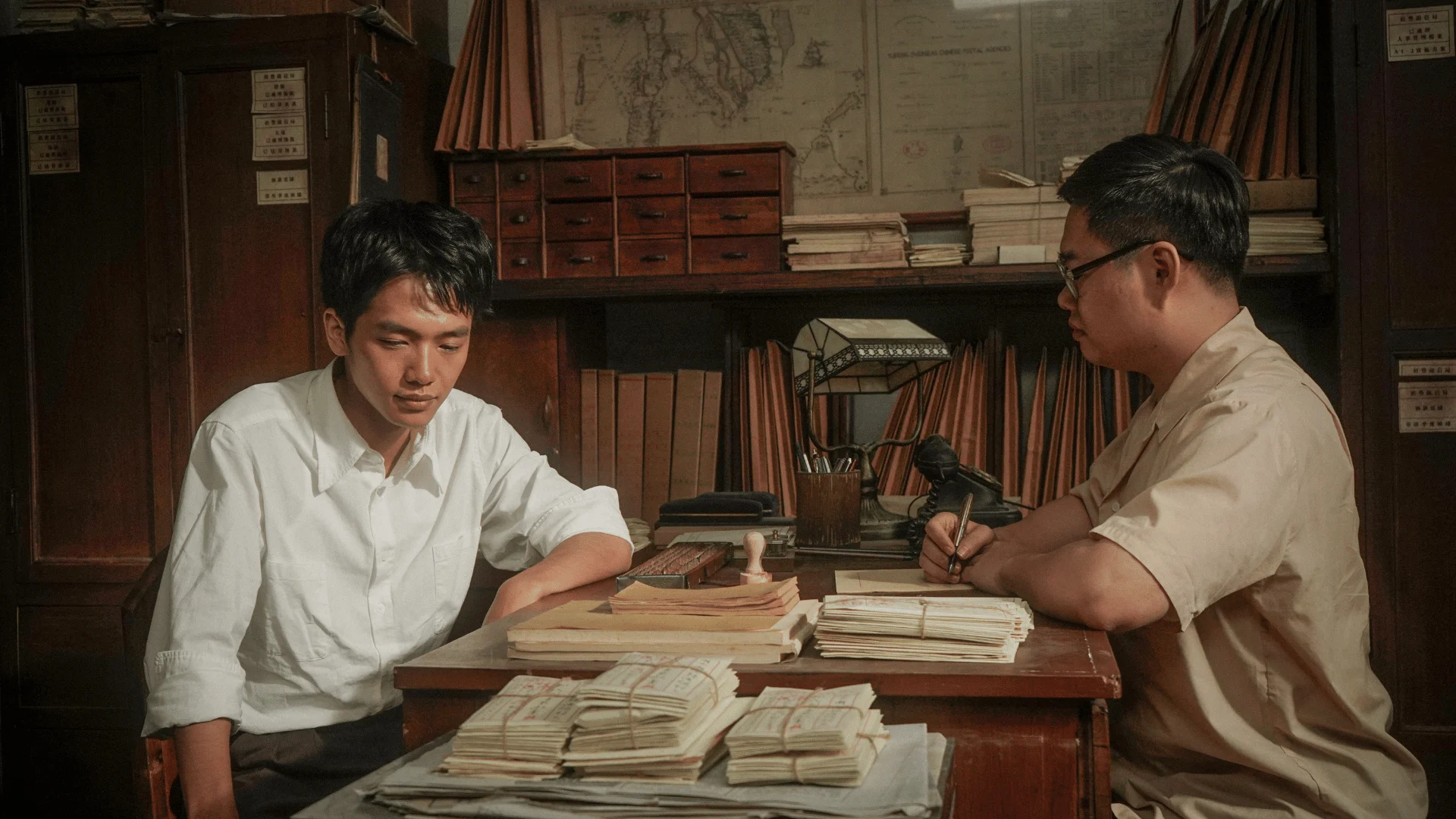 A young man sits across from a clerk among bundled letters, account books, map drawers and stamps in an official Dear You still.