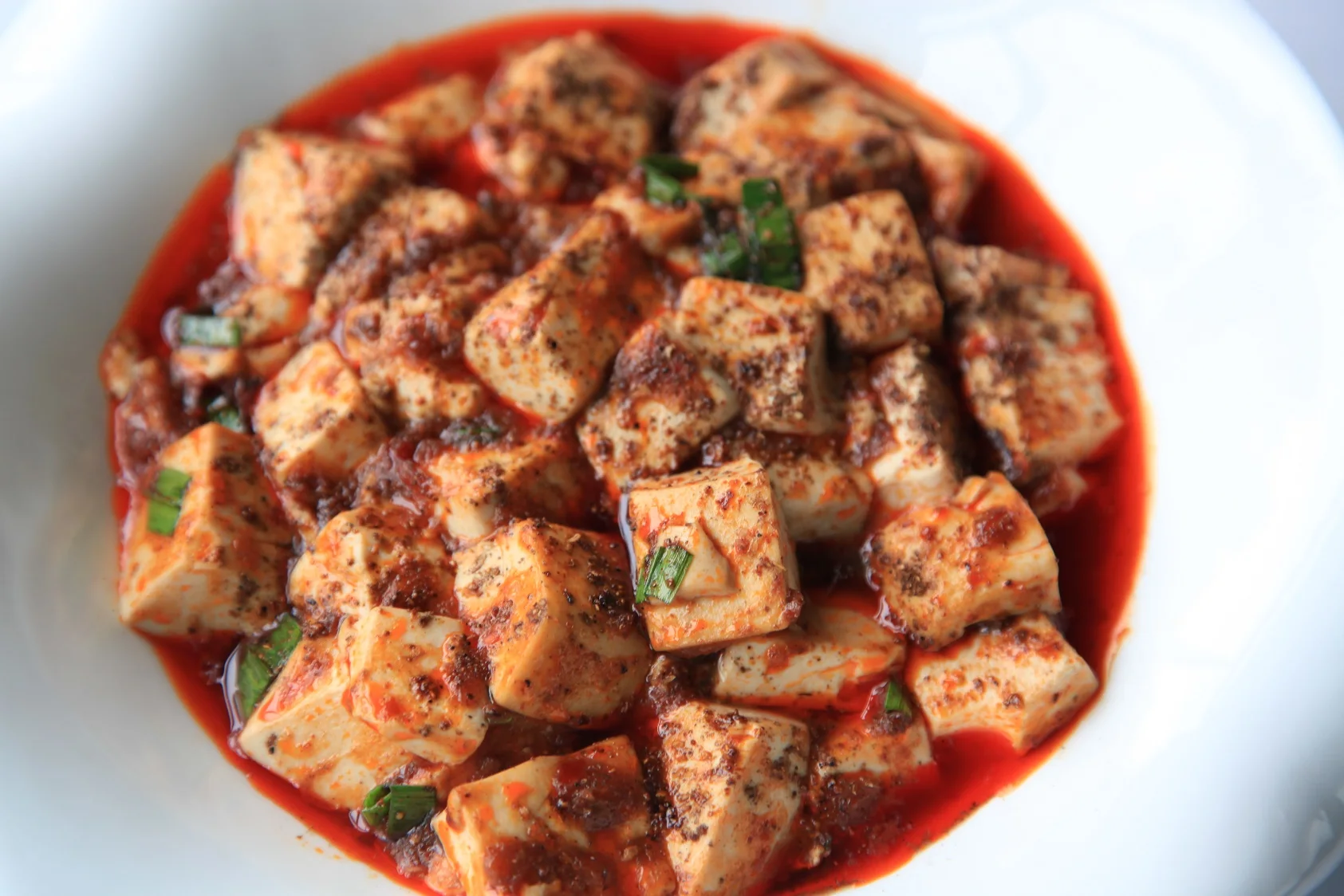 A bowl of mapo tofu with tofu cubes, chilli oil, minced aromatics and a visible dusting of ground Sichuan pepper.