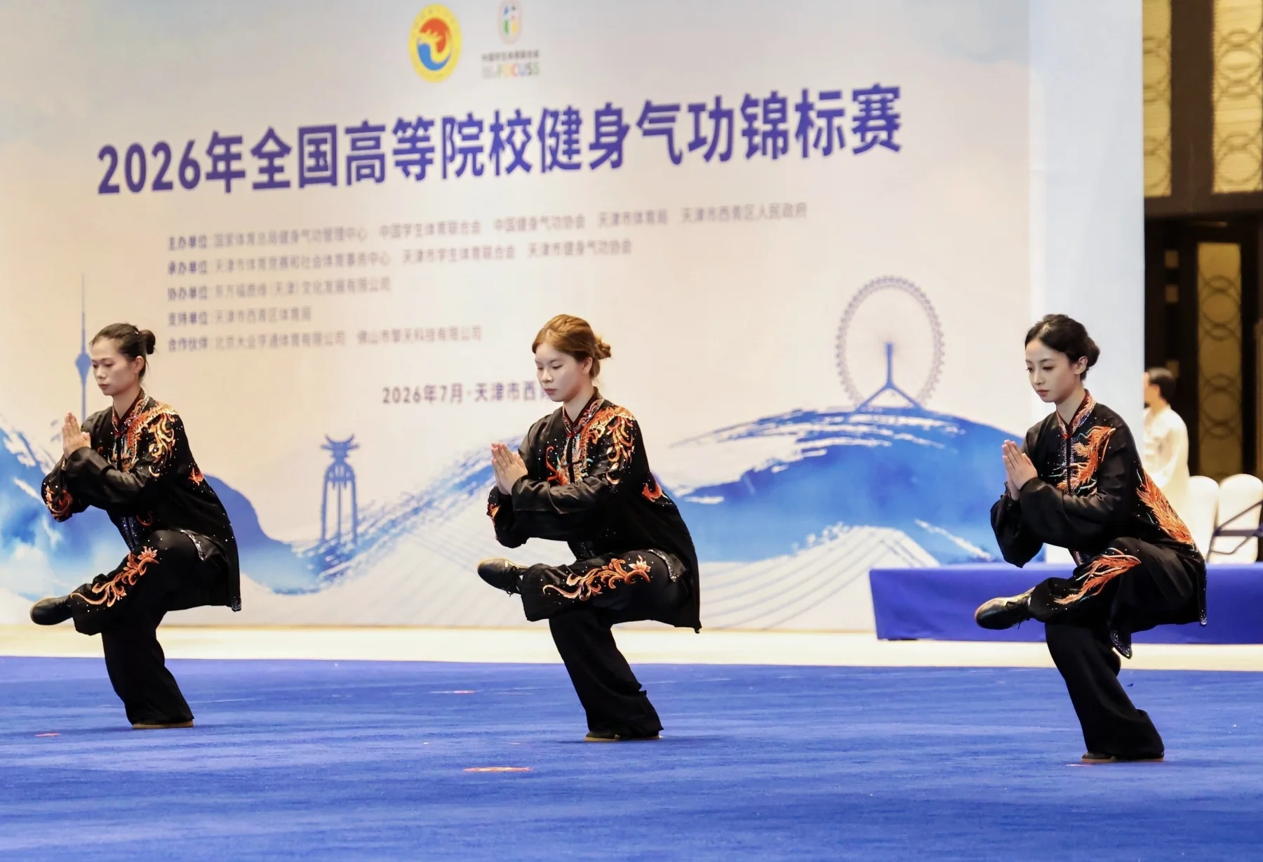 Three young women perform a low balanced Health Qigong movement in coordinated black uniforms at the 2026 national university championship in Tianjin.