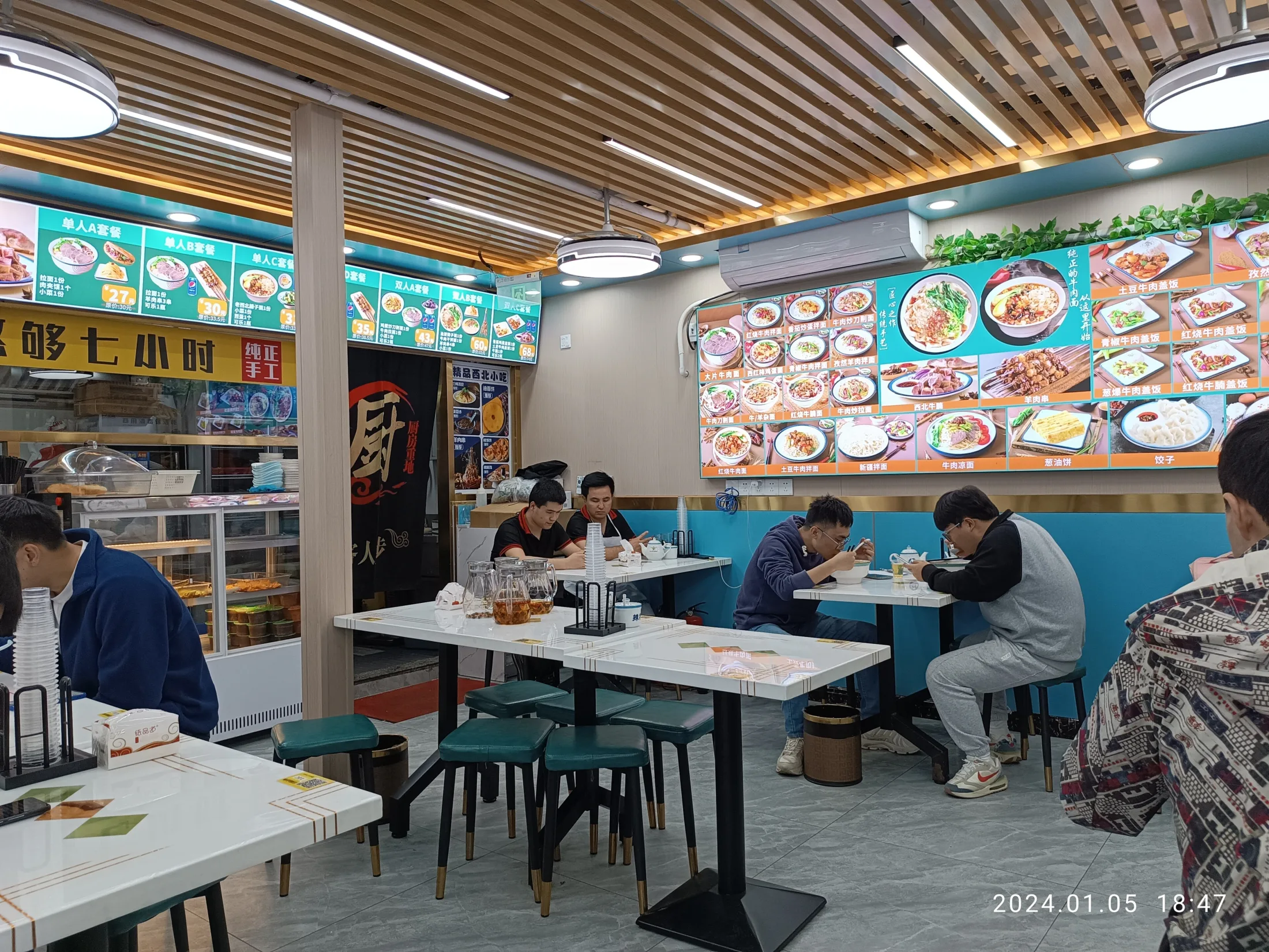 Diners sit in small groups inside a brightly lit Shenzhen noodle restaurant with menu boards covering the wall.