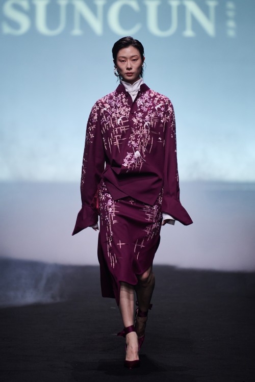 A SUNCUN runway model wears a burgundy high-necked outfit with patterned surfaces, a wrapped waist and asymmetric layered hem.