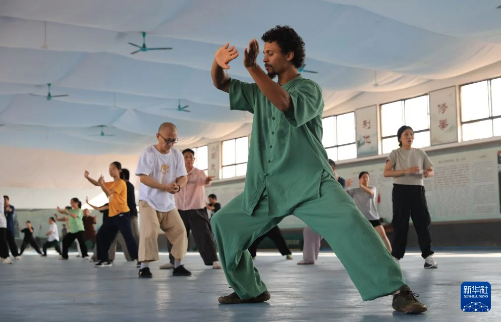 A young international student in green practice clothes trains Chen-style tai chi with a mixed group inside a Chenjiagou martial arts school in July 2026.