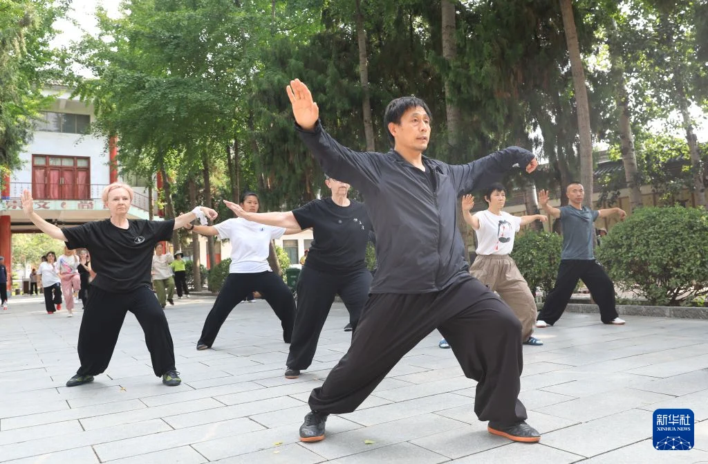 A Chinese tai chi teacher leads international and Chinese students through a wide standing posture outdoors in Chenjiagou in July 2026.
