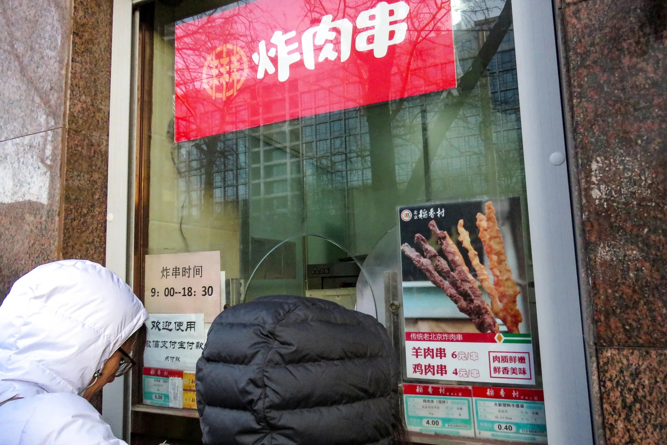 Customers stand at a Beijing fried-kebab window where mobile-payment notices sit beside prices and opening hours.