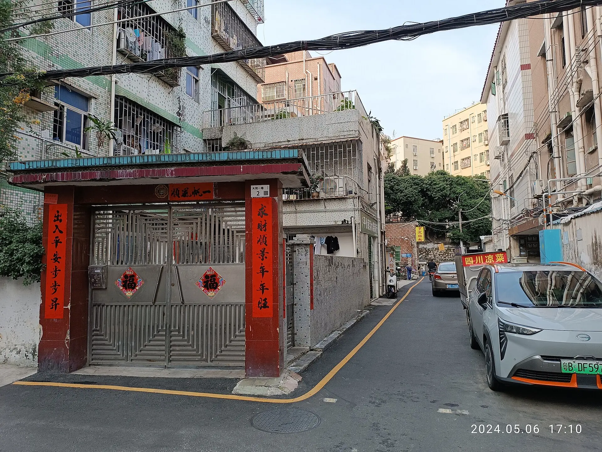 A narrow lane in Bantian, Shenzhen, lined by tiled apartment buildings, gates, overhead cables, small businesses and parked vehicles.