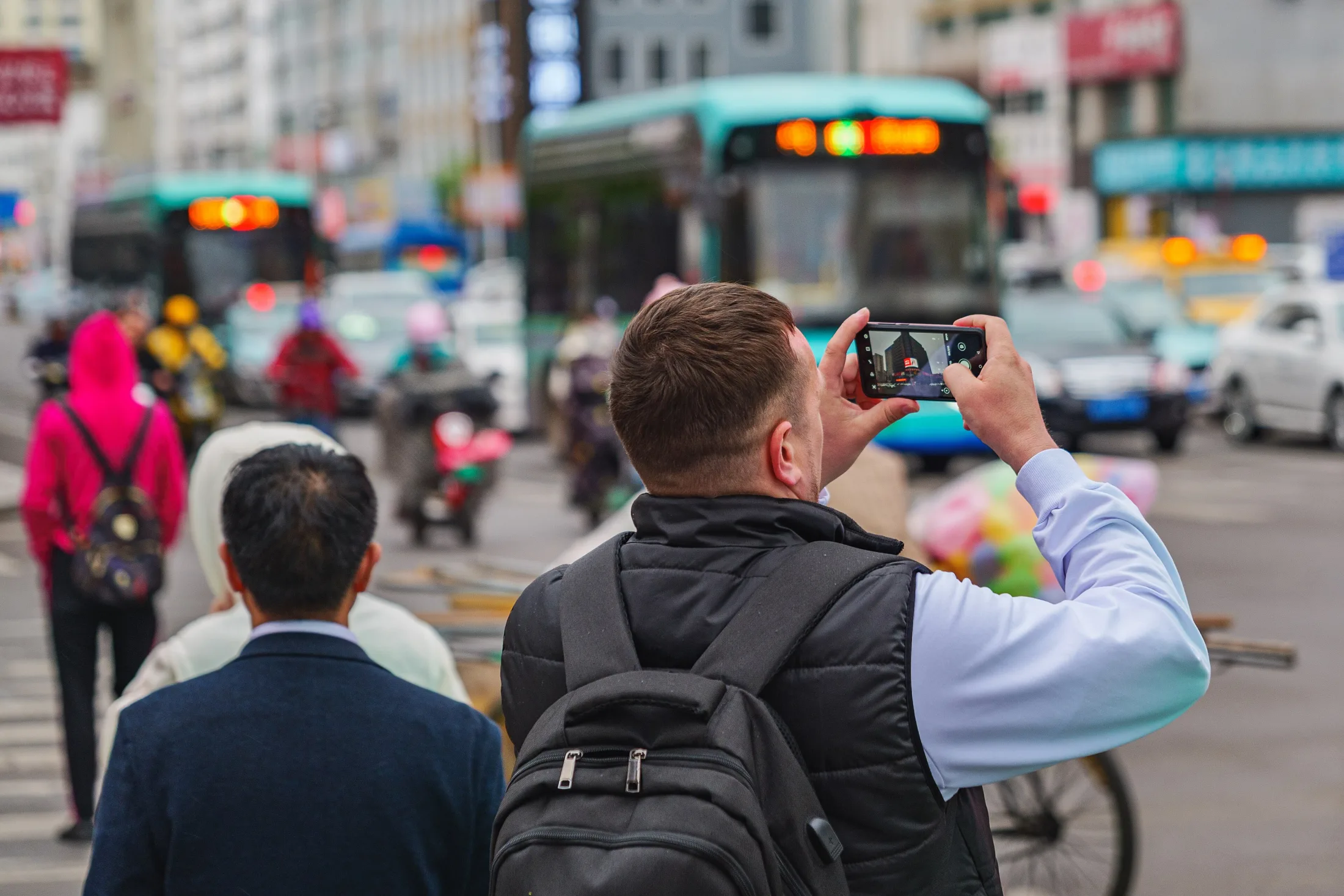 A visitor with a backpack holds up a smartphone on a busy street in Yanji as buses and taxis move behind him.