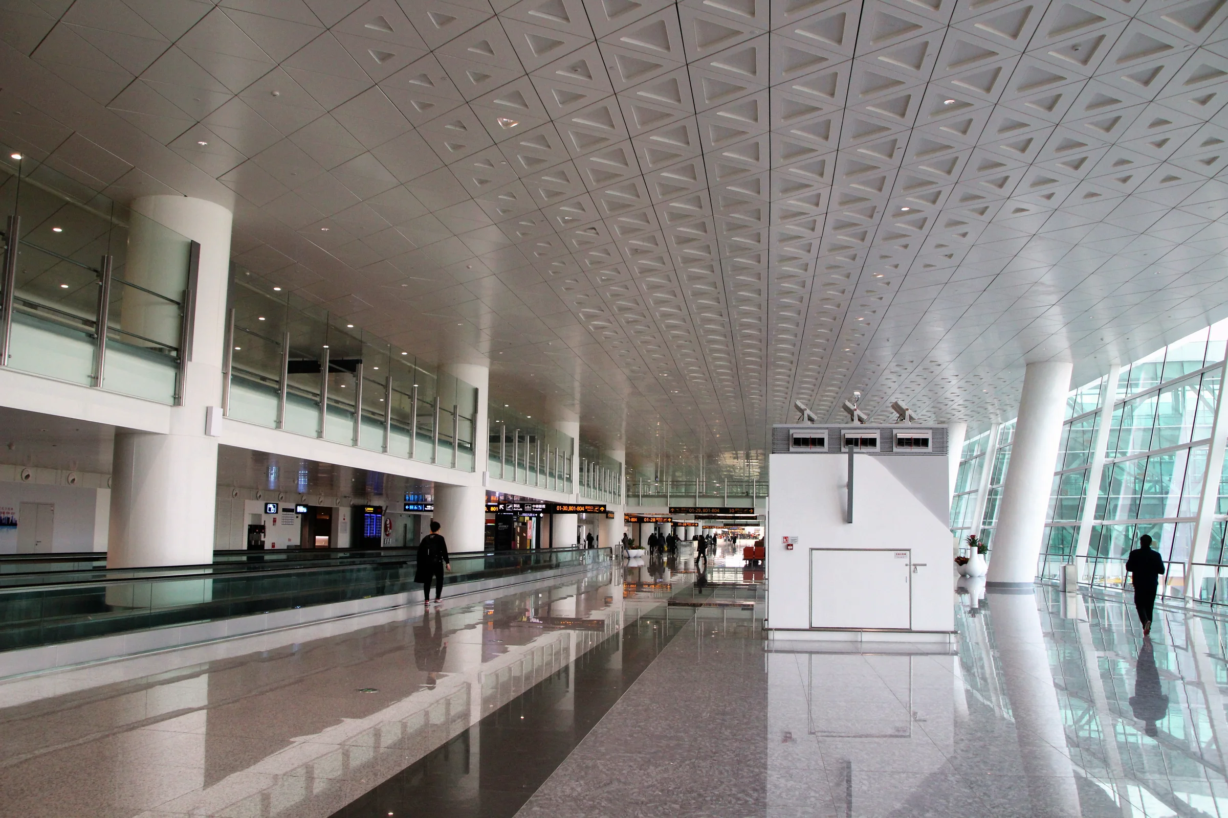 Travellers cross the reflective concourse of Wuhan Tianhe Airport Terminal 3 beneath a long geometric ceiling and bilingual signs.