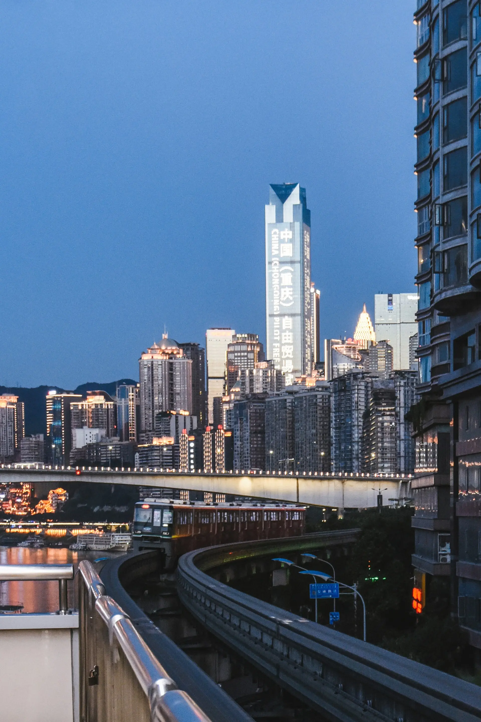 A Chongqing Rail Transit Line 2 monorail curves along the Jialing River between a steep residential slope and the city skyline at dusk.