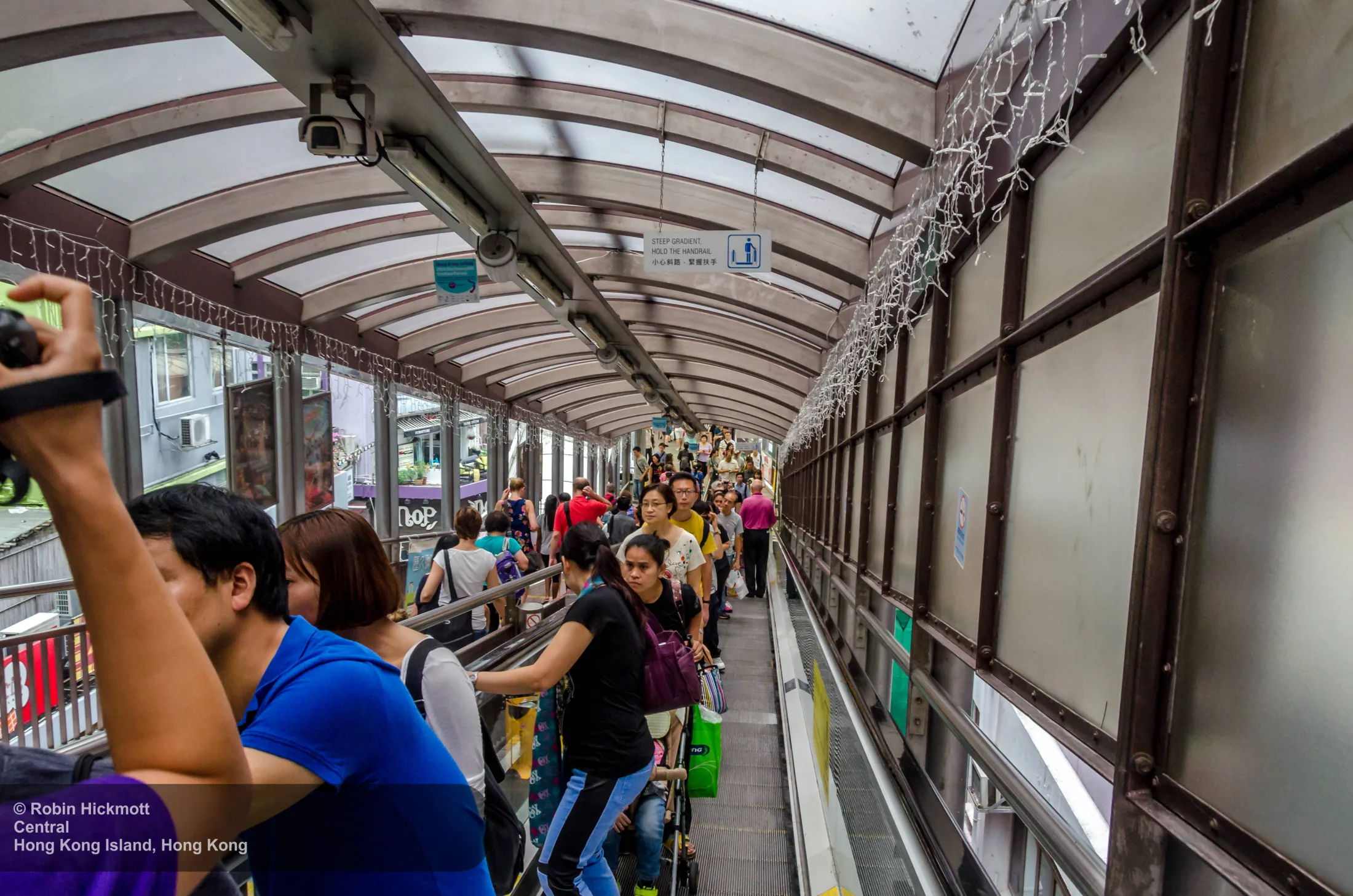 A covered Central–Mid-Levels escalator carries a dense stream of pedestrians through a steep Hong Kong street section.