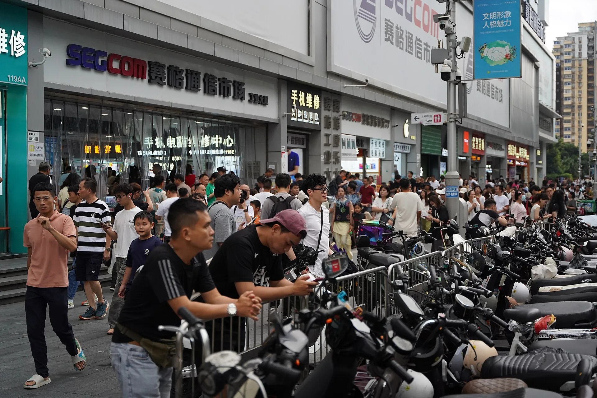 A crowded Huaqiang North Road outside SEG Communications Market, with repair signs, shoppers and parked electric scooters.